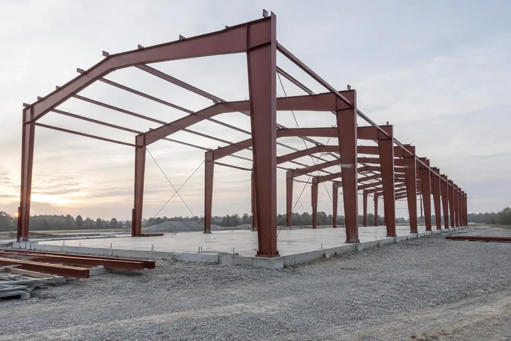Row of red-primed steel rigid frames standing on a concrete slab before roof and wall panels are installed